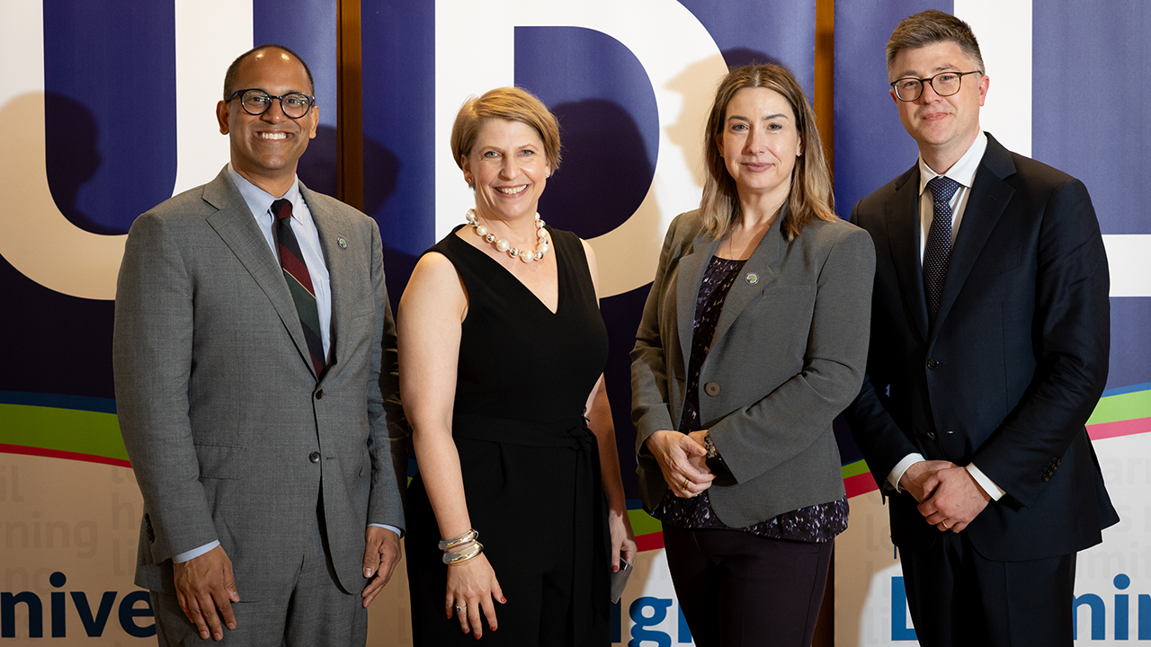 Photo of four people smiling and dressed in professional attire, standing in front of three banners spelling out UDL.